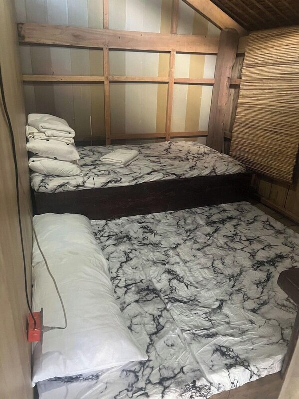 A small, compact room with a bunk bed and a single bed. The room has a wooden ceiling and walls, and there is a window with a bamboo blind.