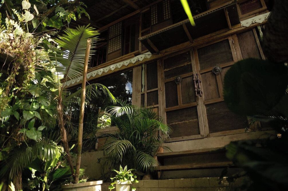 Un edificio tradicional de madera con múltiples ventanas y un balcón, rodeado de abundante vegetación.