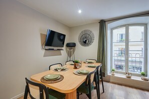 A dining area with a TV mounted on the wall, a decorative mirror, and a window with a view of buildings. - Elegant Apartment with AC - Center of Paris (Paris)