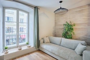 A modern living room with a sofa, a large window with a view of buildings, a hanging light fixture, and a potted plant. - Elegant Apartment with AC - Center of Paris (Paris)