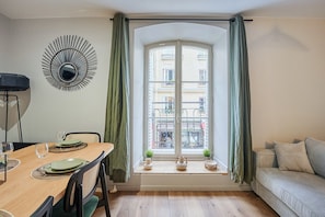 A dining area with a table set for two, a sofa, a window with curtains, and a decorative wall piece. - Elegant Apartment with AC - Center of Paris (Paris)