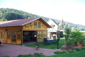A wooden building with a covered patio, a dog on a leash, and a grassy area with potted plants. - Holiday apartment with 2 bedrooms (Bad Schandau)