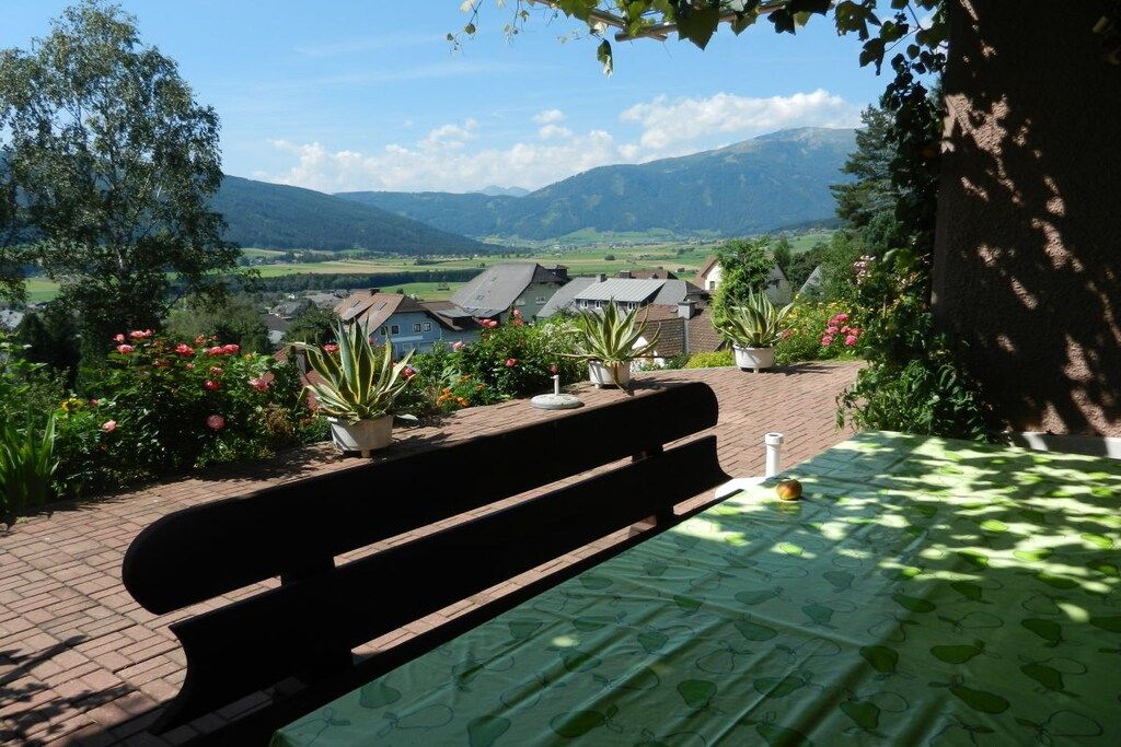 A bench with a table covered by a green tablecloth, surrounded by potted plants and flowers, overlooking a village with houses and mountains in the distance.