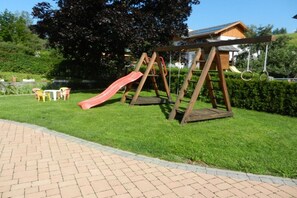 Une aire de jeux avec un toboggan et des balançoires, entourée d’une pelouse bien entretenue et d’un chemin en briques.