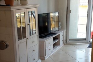 A white entertainment unit with a flat-screen TV, a cabinet with glass doors, and a sliding glass door leading outside. - Holiday apartment directly on the beach (Ostseebad Baabe)