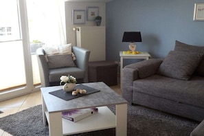 A living room with a grey sofa, a grey armchair, a coffee table with a vase and decorative items, a small side table with a lamp, and a radiator. - Holiday apartment directly on the beach (Ostseebad Baabe)