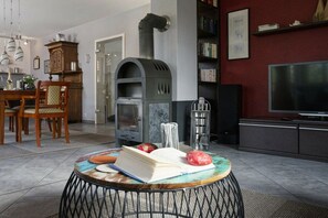 A living room with a fireplace, a dining area with chairs, a bookshelf, and a television. - Holiday home in Glowe with swimming pool (Glowe)