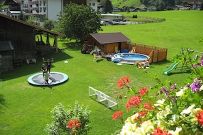 Un espace extérieur verdoyant avec un trampoline, une petite structure en bois et une piscine.