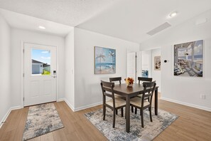 A dining area with a wooden table and chairs, a painting of a beach scene, and a view of the ocean through a door. - Fowl Play - 3/2 w/ 4-Seat Golf Cart in The Villages - Oak Hallow (The Villages)