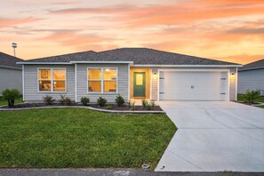 A single-story house with a garage, a well-maintained lawn, and a clear sky at sunset. - Fowl Play - 3/2 w/ 4-Seat Golf Cart in The Villages - Oak Hallow (The Villages)