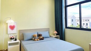 Quarto de hotel com cama, duas almofadas, mesa de cabeceira com luminária, quadro emoldurado e janela com vista para prédios.