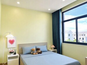 Quarto de hotel com cama, duas almofadas, mesa de cabeceira com luminária, quadro emoldurado e janela com vista para prédios.