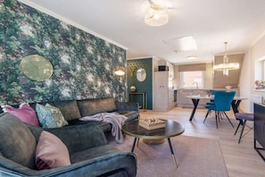 Een moderne woonkamer met een bank, salontafel en een schaakbord. Er is een eethoek met stoelen en een tafel. De kamer heeft een klok aan de muur en een kroonluchter.