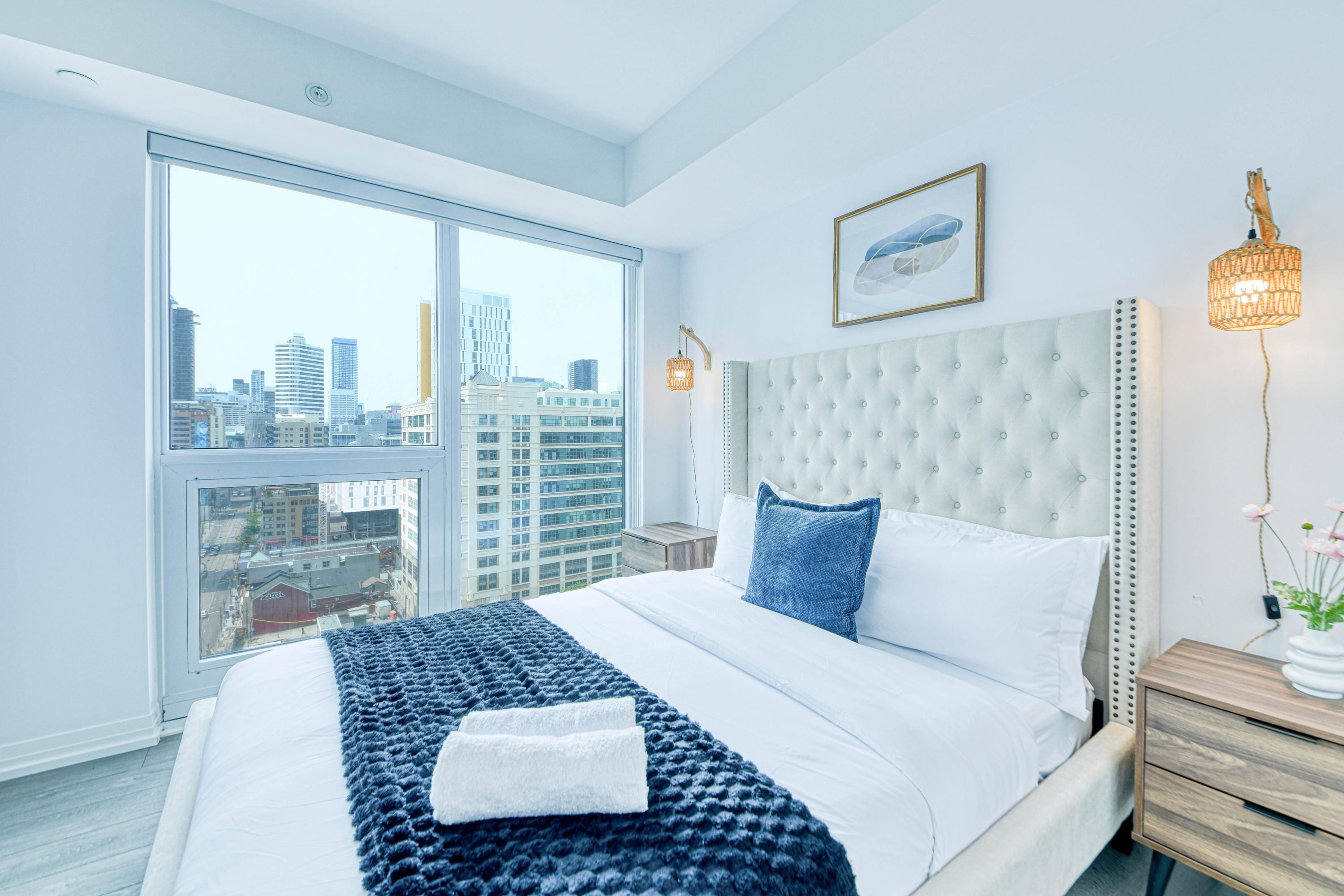 A modern bedroom with a large bed, a city view, and a cityscape visible through the window.