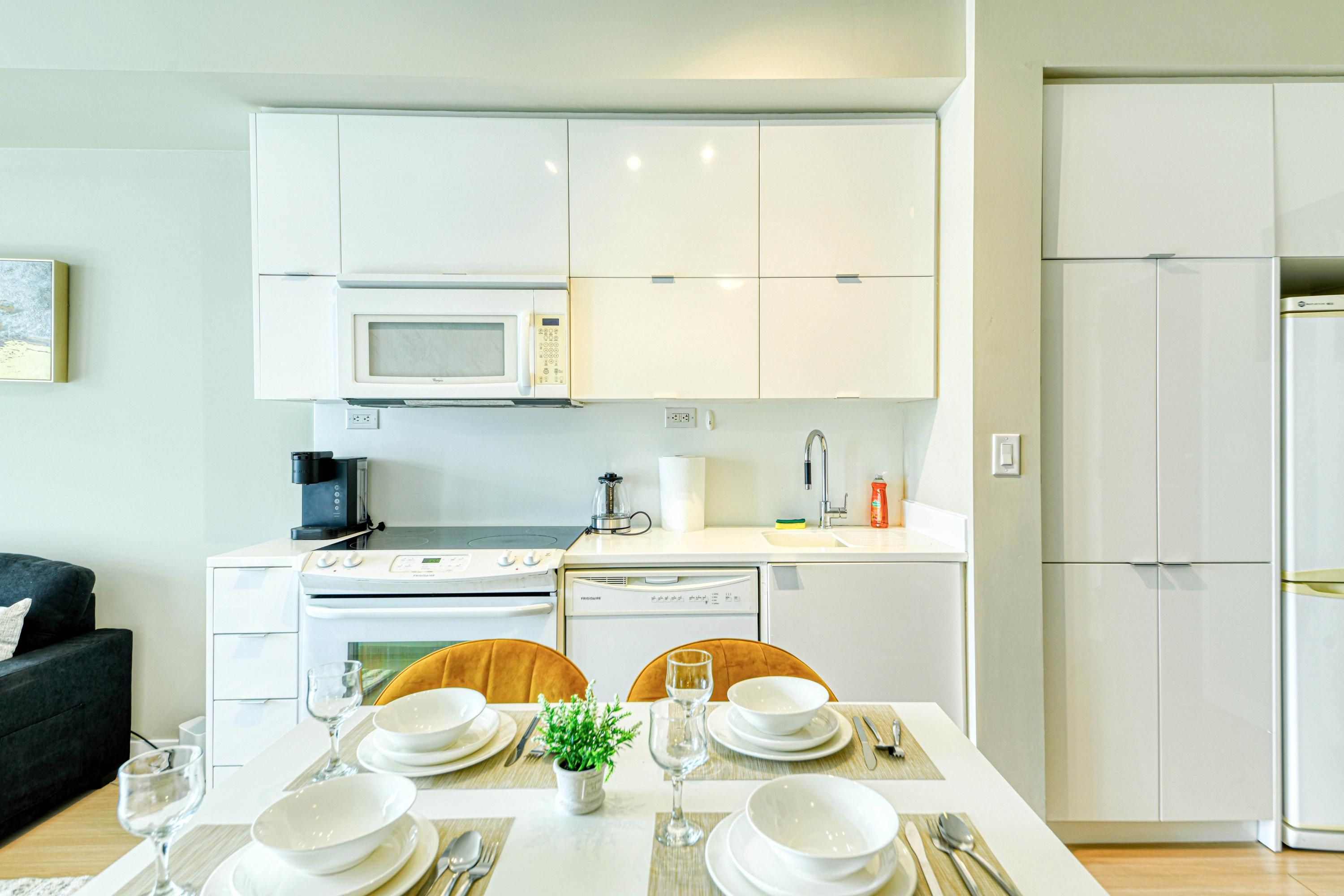 A modern kitchen with white cabinets, a microwave, a stove, and a dishwasher. There is a dining table set with plates, glasses, and cutlery.