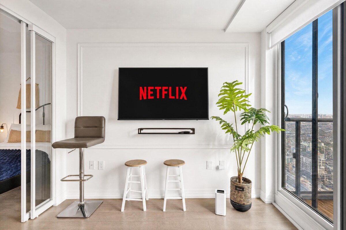 Un salón moderno con una gran pantalla de televisión mostrando 'NETFLIX', un sofá, dos taburetes altos, una planta en maceta y vistas de la ciudad a través de un ventanal.