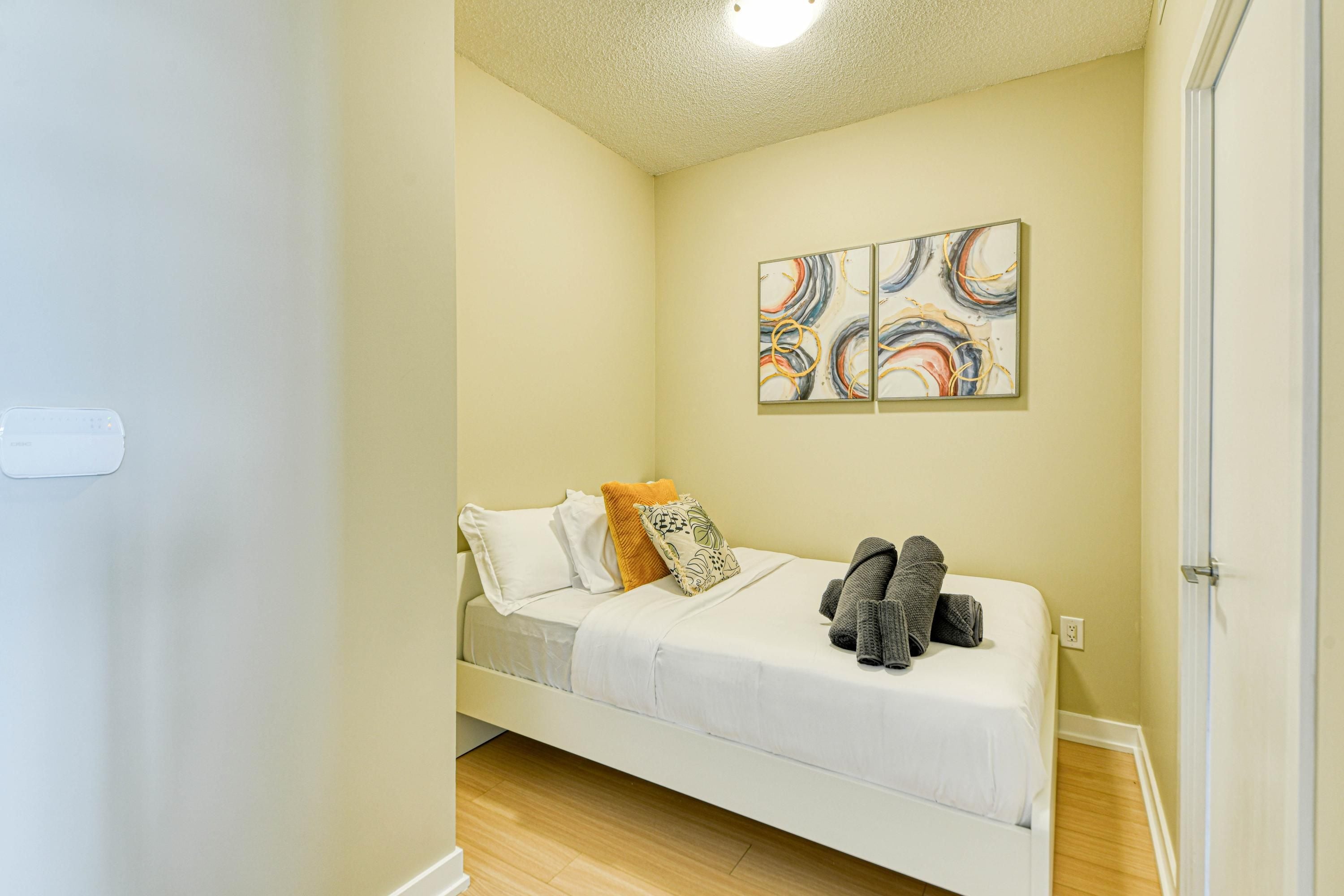 A small, single-bed room with a wooden floor, a white bed, and a wall-mounted abstract art piece.