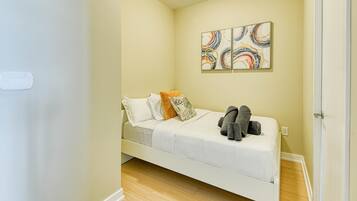 A small, single-bed room with a wooden floor, a white bed, and a wall-mounted abstract art piece.