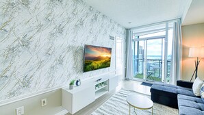 A modern living room with a large TV on a white entertainment unit, a grey sofa, and a view of a balcony with outdoor furniture. - StayToronto - Terrace 21 Apartments (Toronto)