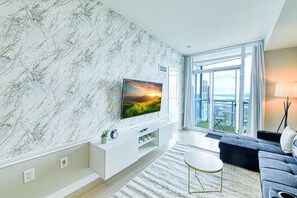 Uma sala de estar moderna com uma televisão grande em uma unidade de entretenimento branca, um sofá cinza e vista para uma varanda com mobiliário de exterior.