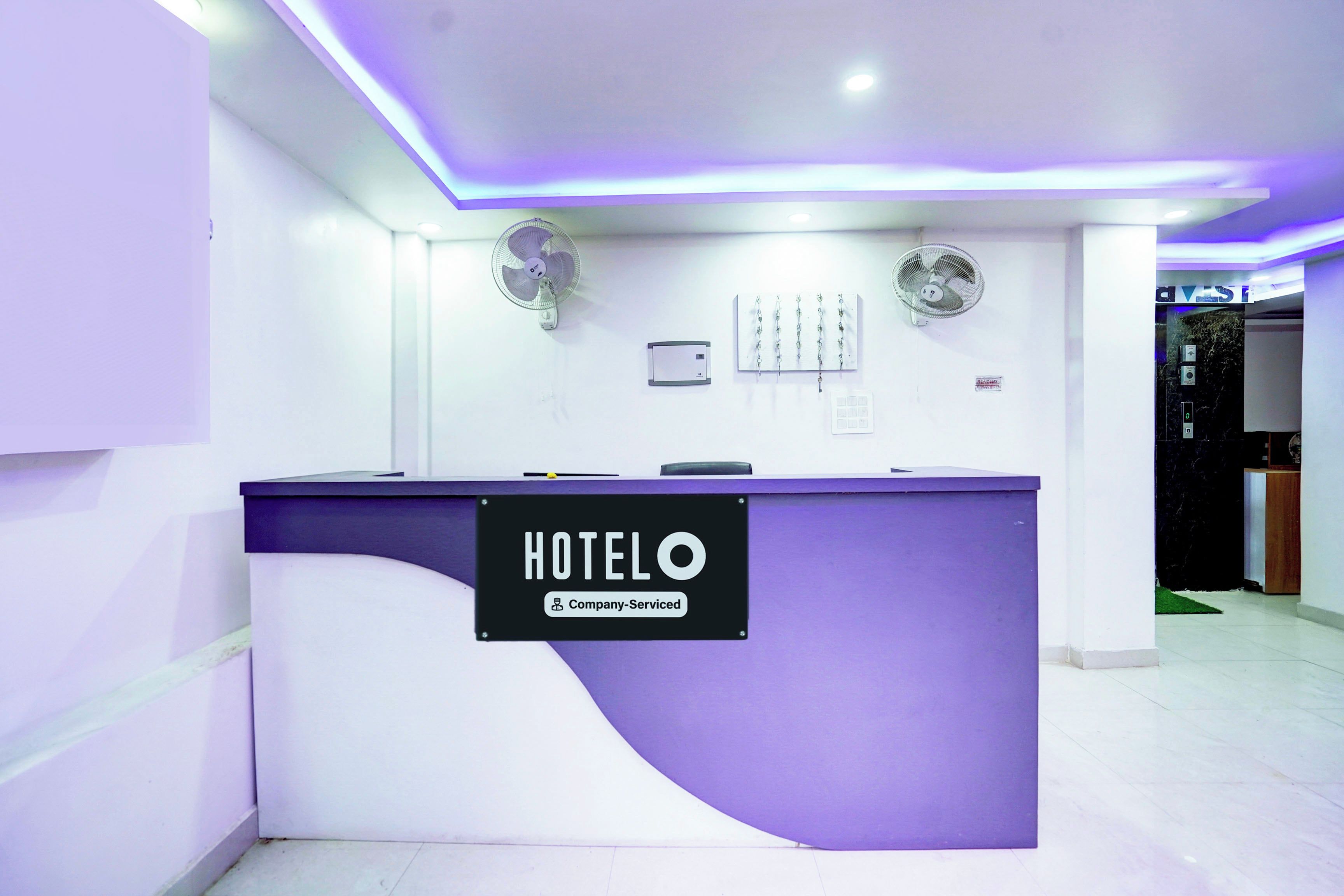 Un mostrador de recepción de hotel con un letrero que indica servicio corporativo. Hay dos ventiladores de pared y una pantalla digital encima del mostrador.