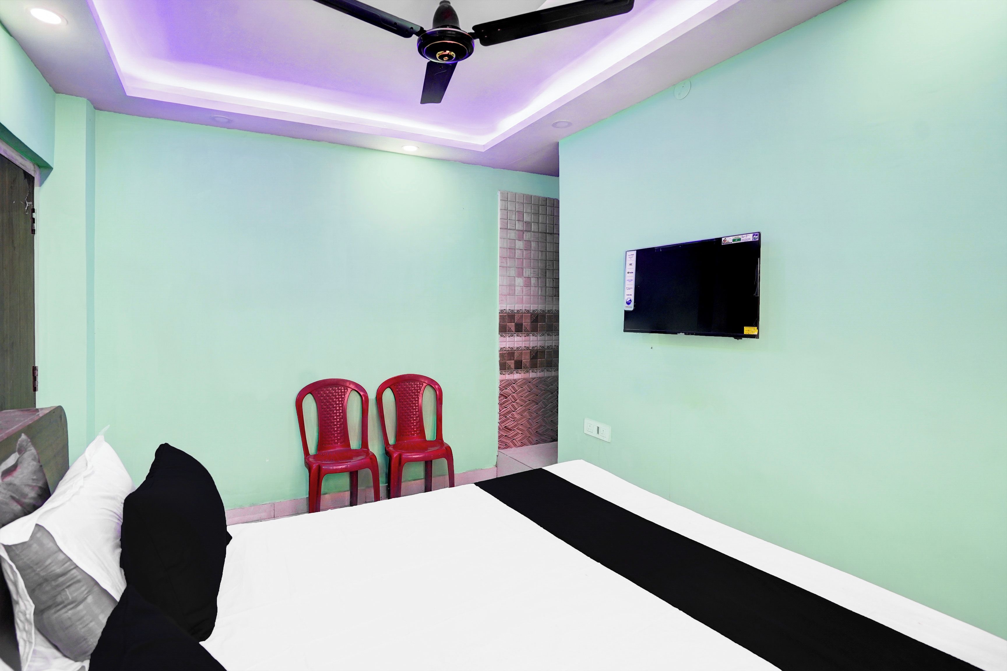Un dormitorio con una cama, dos sillas rojas, un televisor en la pared y un ventilador de techo.