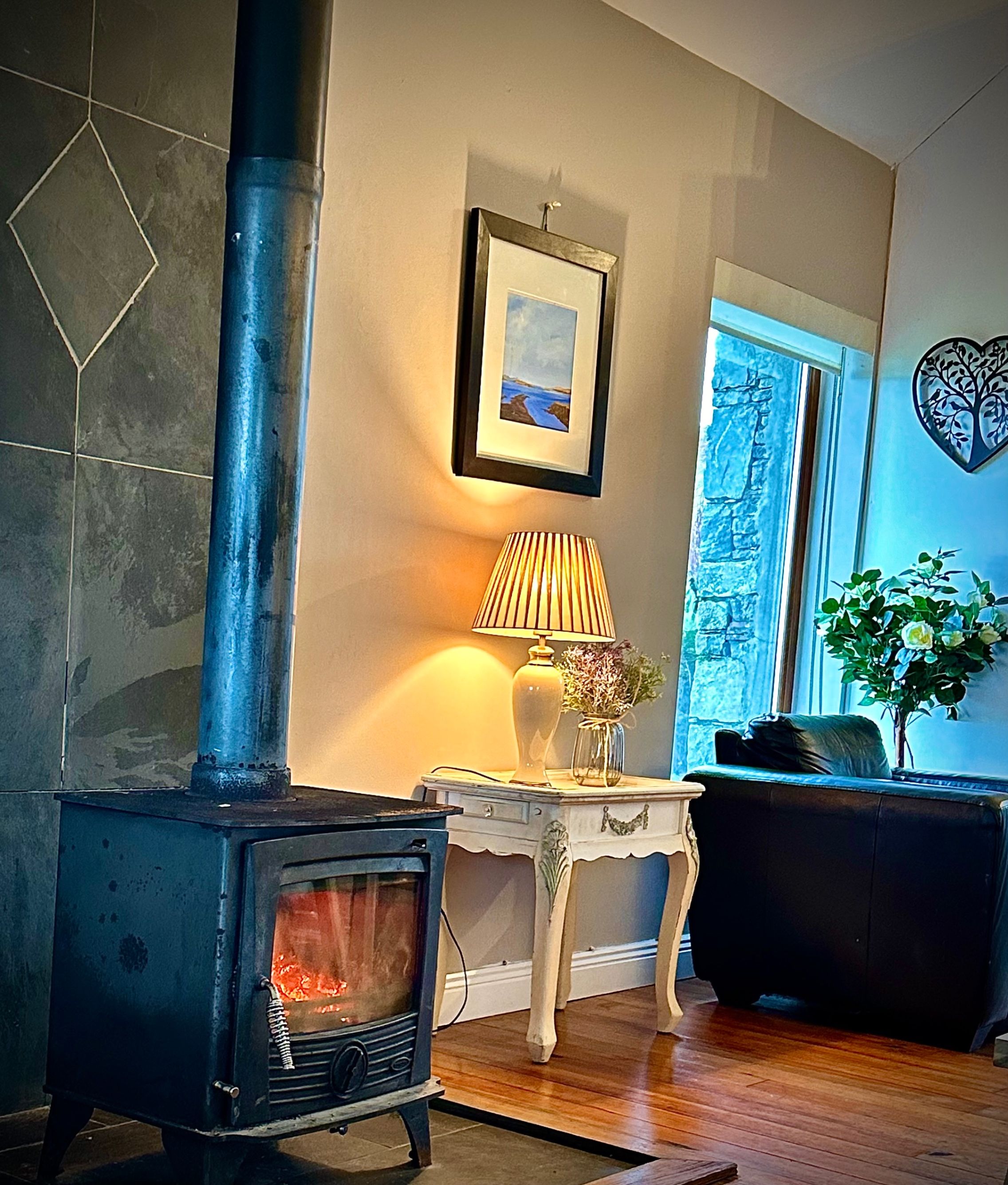 A room with a wood-burning stove, a framed picture on the wall, a lamp on a small table, and a window with a view.