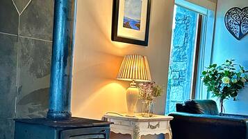 A room with a wood-burning stove, a framed picture on the wall, a lamp on a small table, and a window with a view.
