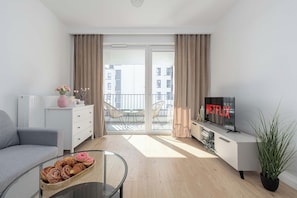 Eine moderne Wohnung mit einer Couch, einem Glastisch mit Gebäck, einem Fernseher mit 'NETFLIX' auf dem Bildschirm und einem Balkon mit Blick auf Gebäude.