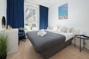 Ein Hotelzimmer mit Bett, Schreibtisch, Stuhl, einem Fenster mit Vorhängen, einem Bild an der Wand und einer Pflanze.