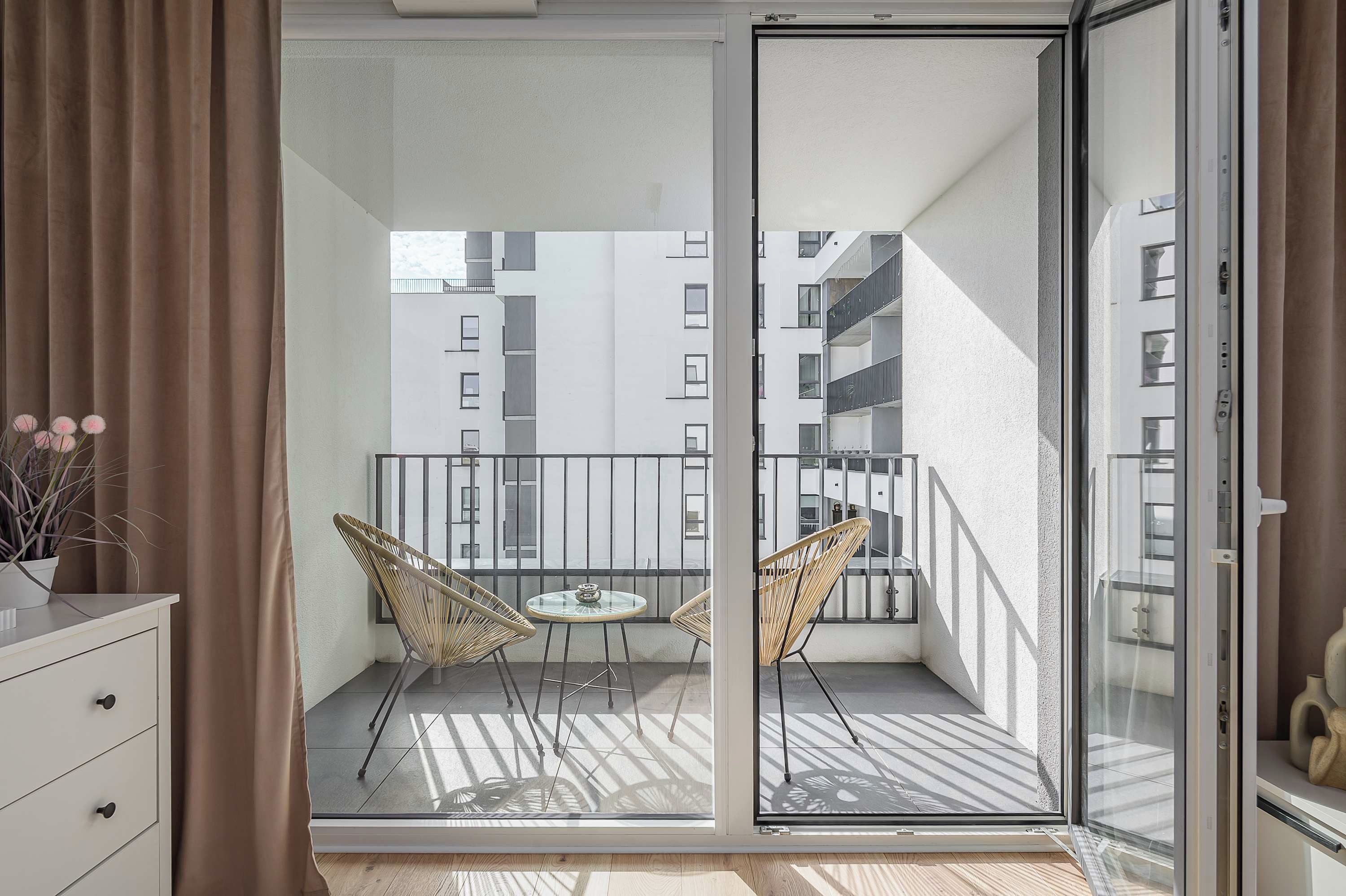 A balcony with two wicker chairs and a small table, overlooking a building with multiple windows.
