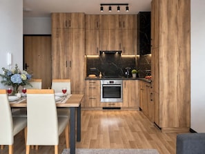 A modern kitchen with wooden cabinets, a built-in oven, and a dining area with a table and chairs. - Lumina 2-room Apart w/ Parking I Balcony (Warszawa)