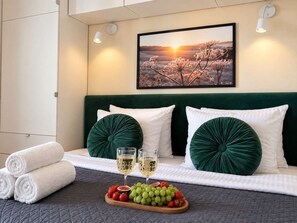 Ein ordentlich bezogenes Bett mit grünen Kissen, ein Obsttablett und Weingläser sowie ein Sonnenuntergangsgemälde an der Wand.