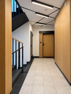 Un couloir moderne avec une porte en bois portant le numéro « 94 », un escalier avec des rampes noires et un plafond doté de luminaires linéaires.