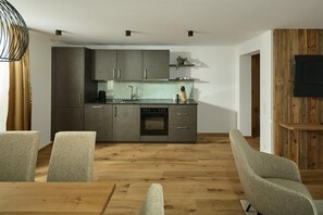 A modern kitchen with grey cabinets, a built-in oven, and a sink. There are two grey chairs and a wooden dining table in the foreground. - Modern suite with outdoor sauna (Gaschurn)