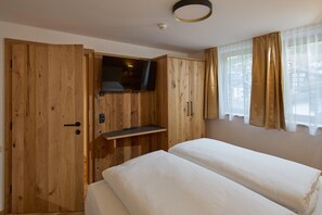 A room with a wooden wardrobe, a television mounted on the wardrobe, a bed with white bedding, and a window with sheer curtains. - Modern suite with outdoor sauna (Gaschurn)