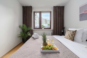 Ett hotellrum med en säng, ett fönster med gardiner, ett element, en växt och ett bricka med frukt och glas.