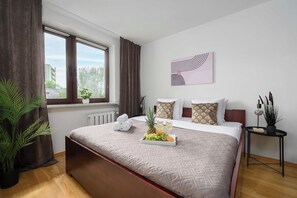 Ein Hotelzimmer mit einem großen Bett, einem fenster mit Vorhängen, einem Heizkörper, einer kleinen Pflanze und einem an der Wand hängenden Kunstwerk.