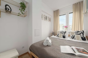 Ett hotellrum med en säng, ett fönster med gardiner, en växt som hänger på väggen och en inramad tavla.