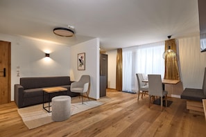Una sala de estar moderna con un sofá gris, dos sillones, una mesa de centro de madera y un pequeño reposapiés. Hay un comedor con una mesa y sillas, y un ventanal con cortinas.