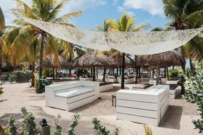 Ein Strandloungebereich mit Reetdachschirmen, weißen gepolsterten Sitzgelegenheiten und freiem Blick auf das Meer.