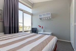 Ein Hotelzimmer mit einem großen Bett, einem Sessel, einem kleinen Tisch mit Blumen und einem Vorhangfenster sowie einem an der Wand befestigten Regal.