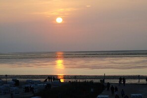 A beach scene with the sun setting over the water, people walking along the shore, and beach huts in the foreground. - Holiday apartment with 2 bedrooms (Norden)