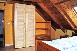 Een kamer met een schuine balkenplafond, een bed met een blauw beddegoed met patroon en een grote houten garderobe.
