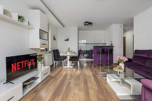 Un salon moderne doté d’un téléviseur à écran plat, d’un coin repas avec une table et des chaises, d’une cuisine aux meubles violets et d’un canapé violet.