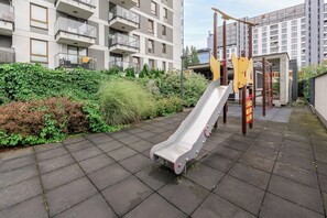 Een speeltuin met een glijbaan en klimconstructies, omringd door een haag en een gebouw met balkons.