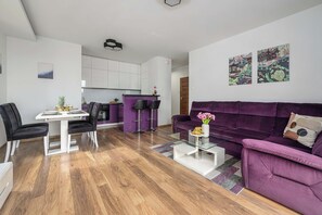 A modern living room with a purple sofa, a dining area with a white table, and a kitchen with white cabinets. - Lumina 3-Room Apt with Balcony & City View (Warszawa)