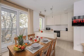A modern kitchen with a dining area, a TV screen displaying 'NETFLIX', and a view of trees through the glass door. - Lumina Spacious 4 Room Apartment with Parking (Warszawa)