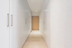 A hallway with white walls, a wooden door at the end, and a light fixture on the ceiling. - Lumina Spacious 4 Room Apartment with Parking (Warszawa)