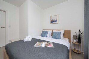 Cama bem arrumada com colcha cinza acolchoada, um par de toalhas enroladas e um livro aberto sobre a cama.